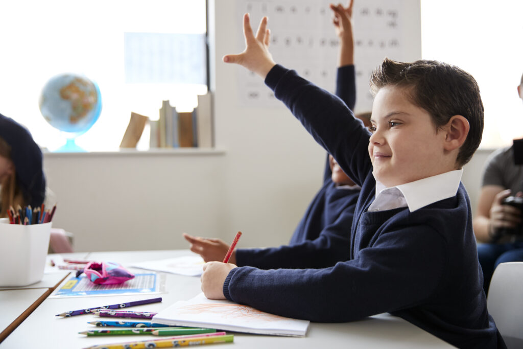 Student with special learning needs sitting at a desk raising his hand in a primary school class, close up, side view